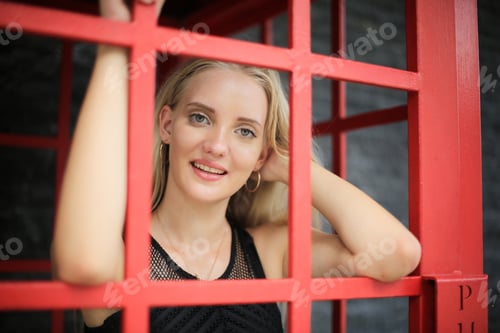 Preview: Portrait of A beautiful blonde in a black dress stands in a red phone booth.