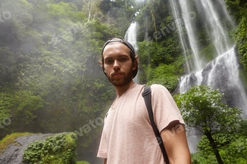 Preview: Handsome young Caucasian male backpacker wearing t-shirt and baseball cap backwards looking down at