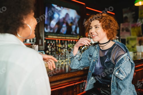 Preview: Young redhead woman talking to blurred african american friend near stand in bar