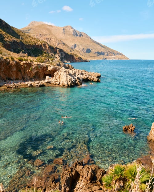 Preview: Coastal view of a rocky shoreline with clear waters and distant mountains under a bright blue sky.