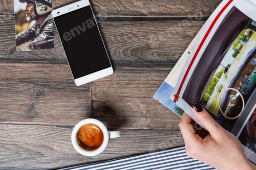Preview: Top view of table with cup of coffe and smartphone. Hand holding magazine.