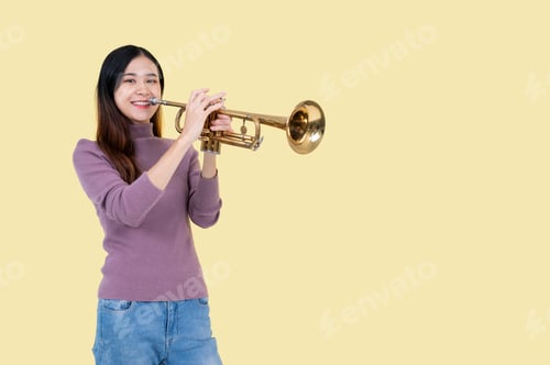 Preview: A talented Asian woman is playing a trumpet while standing against an isolated yellow background.
