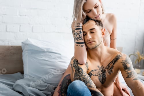 Preview: Sexy woman in bra embracing shirtless tattooed boyfriend on bed