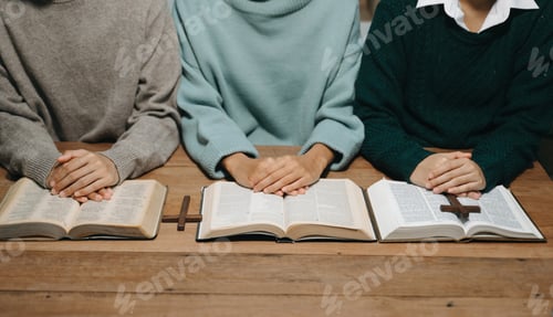 Preview: Group of Christians sit together and pray around a wooden table with blurred open Bible pages