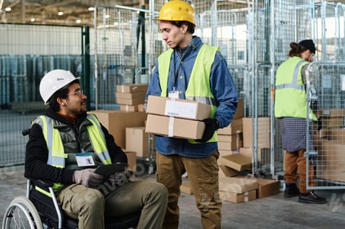 Preview: Young engineer with boxes talking to colleague in wheelchair using tablet