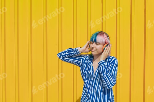 Preview: Beautiful young female with headphones enjoying listening to music standing against a yellow wall