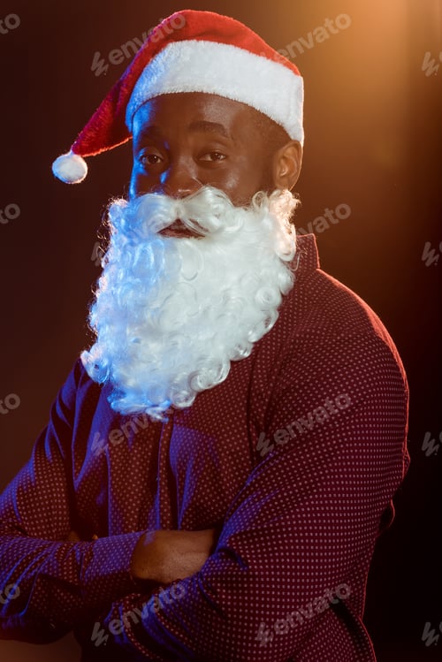 Preview: african american man in santa hat and beard posing with crossed arms on black with backlit