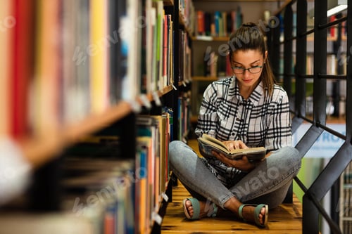 Preview: Young female student study in the library reading book while sitting near bookshelf.
