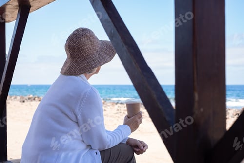 Preview: Back view of senior woman in casual clothing with hat sitting relaxed at the beach looking away