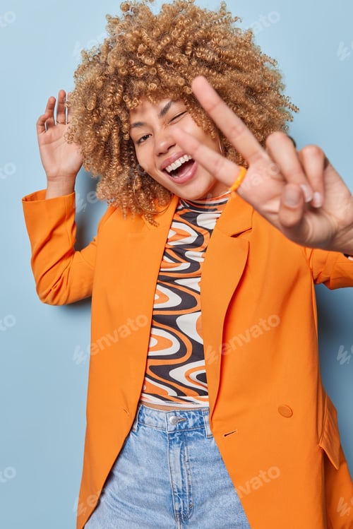 Preview: Vertical shot of positive curly haired young woman wears stylish orange jacket and jeans shows peace