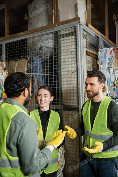 Preview: Smiling male worker in safety vest and gloves talking to indian colleague while resting near waste