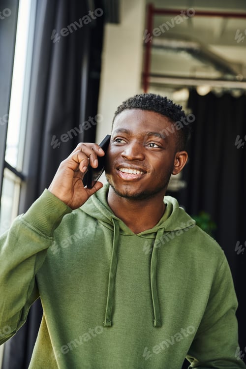 Preview: optimistic african american man in green hoodie talking on mobile phone and looking away in