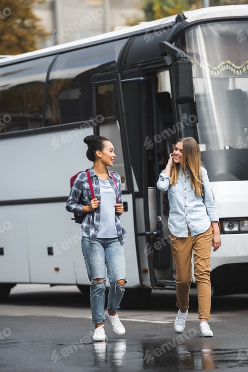 Preview: selective focus of multicultural women with backpacks walking near travel bus at urban street