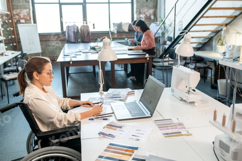 Preview: Young serious designer in wheelchair drawing sketches for new seasonal collection in workshop