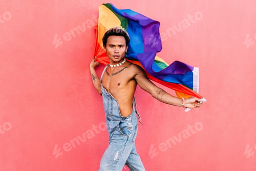 Preview: Young stylish man holding a waving LGBT flag against pink wall outdoors and looking at camera