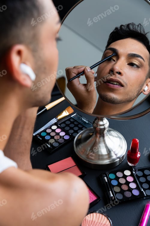 Preview: Biracial transgender man looking in mirror and putting on make-up, applying eyeshadow