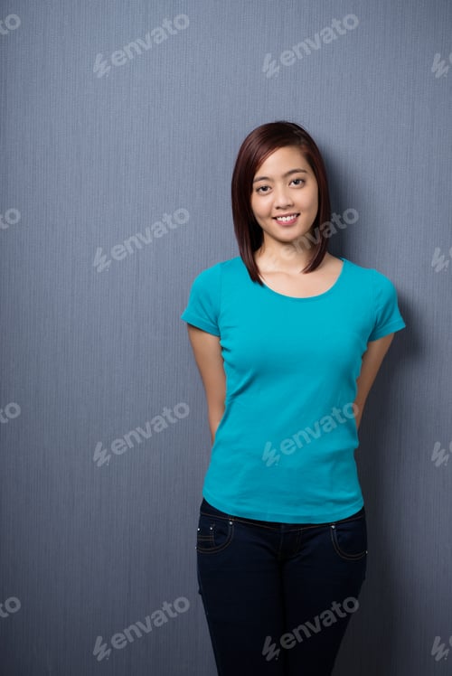 Preview: Attractive Demure Young Asian Woman Standing With Her Hands Behind Her Back Against A Dark Studio