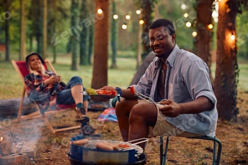 Preview: Young black man grilling food while camping with girlfriend in the woods.