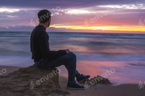 Preview: Man sitting on beach rocks looking at ocean during sunset