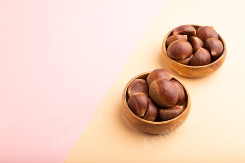 Preview: wooden bowl with raw edible Сhestnuts on pink and orange paper pastel, side view, copy space