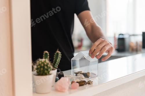 Preview: Crop guy throwing coin into money box in modern apartment