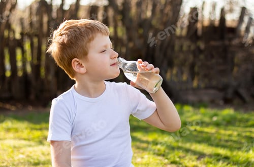Preview: Red-haired teenager boy sitting on the grass at sunset, resting during yoga, drinking water
