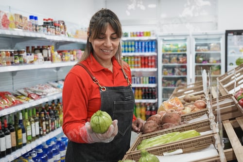 Preview: Mature latin woman working inside supermarket while holding fresh oragnic zucchini