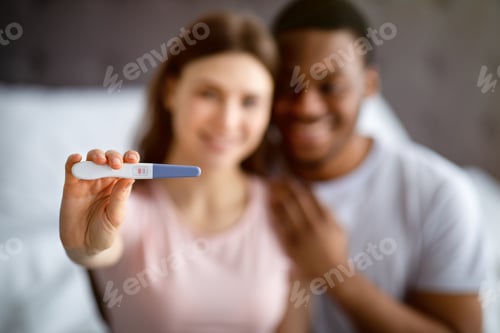 Preview: Happy interracial couple showing positive pregnancy test at home, selective focus, free space