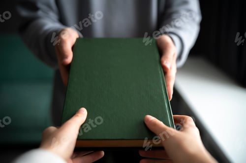 Preview: Two Individuals Exchange a Green Hardcover Book Indoors, giving new knowledge
