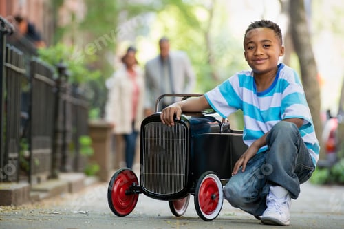 Preview: A young boy playing with a old fashioned toy car on wheels on a city street. A couple looking on.