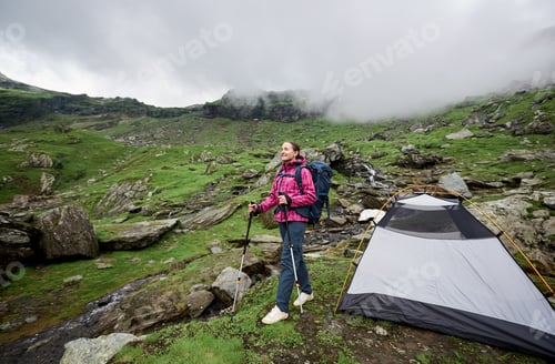 Preview: tourist posing near tent on green grassy rocky meadow with beautiful mountains covered with fog