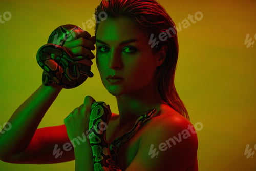 Preview: Stunning woman in a mix of red and green light, holding two pythons in her hands.