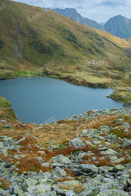Preview: Scenic view of a mountain lake surrounded by rocky terrain and grassy hills under a cloudy sky