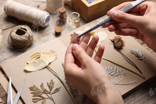 Preview: Woman putting dry flowers into notebook at wooden table, closeup