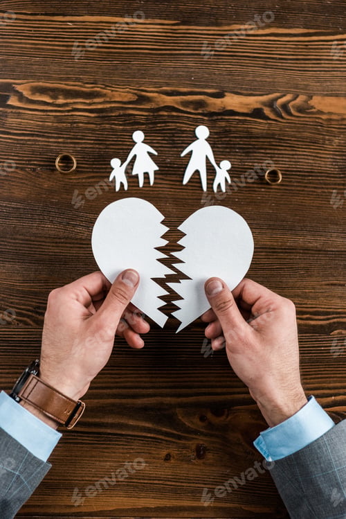 Preview: cropped shot of man holding teared heart with family and engagement rings on wooden table, divorce