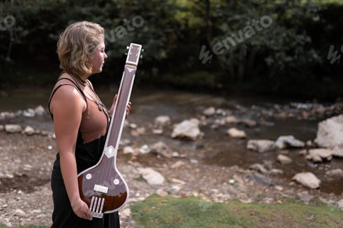 Preview: A young adult woman is holding her indian tanpura next to a river