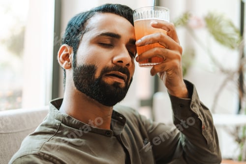 Preview: Sad, tired Indian man feeling sick holding glass of water on forehead. Health care concept