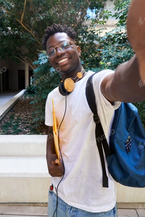 Preview: Vertical selfie of student male wearing headphones and holding a file folder at campus.