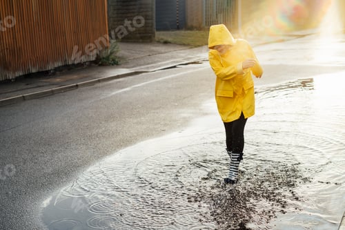 Preview: Woman having fun on the street after the rain. Smiling woman wearing rain rubber boots and raincoat