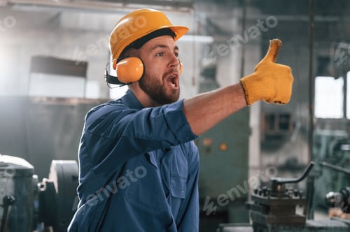 Preview: Man in hard hat and headphones is showing thumb up. Factory worker in blue uniform is indoors