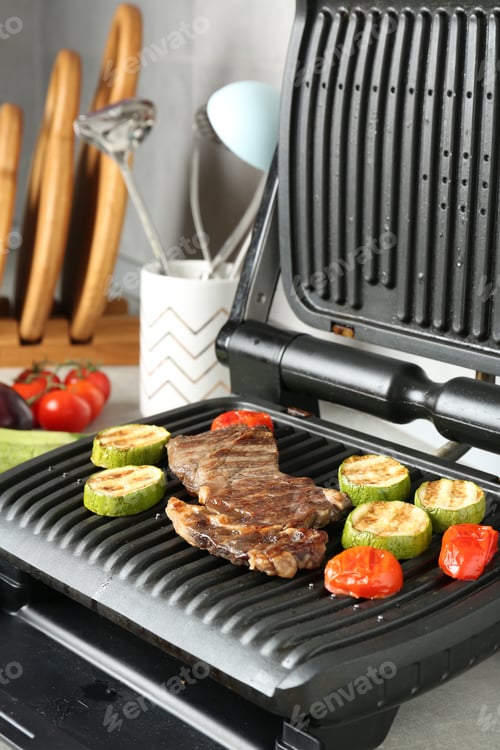 Preview: Open electric grill with beef steak, zucchini and tomatoes on light grey table, closeup