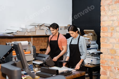 Preview: cheerful man in apron working with printer next to pretty colleague in print center
