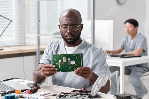 Preview: Serious African man in workwear looking at motherboard in hands
