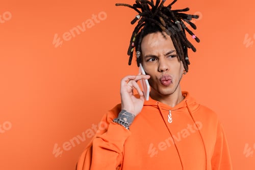 Preview: tattooed multiracial man with dreadlocks pouting lips and talking on cellphone isolated on coral