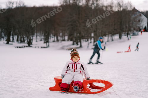 Preview: Little girl sits on a sled on a snowy plain against the backdrop of sledgers and skiers