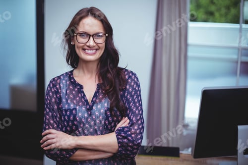 Preview: Woman standing with arms crossed at office desk in front of computer monitor wearing eyeglasses