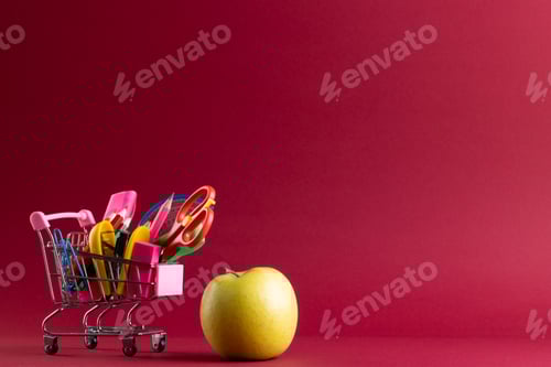 Visualização: Vista de perto do carrinho de compras em miniatura com materiais escolares, maçã e espaço de cópia em fundo vermelho