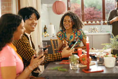 Preview: Group of friends sitting around festive dinner table at home, eating and use mobile phone