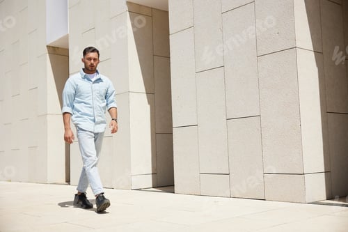 Preview: Confident young man walking outdoors in urban environment with modern architecture, casual attire