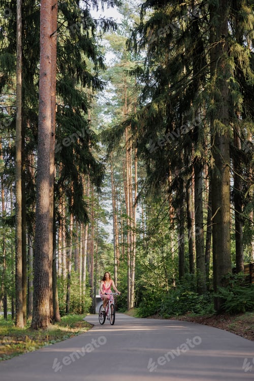 Preview: young active woman in pink dress rides a bicycle in park among the trees